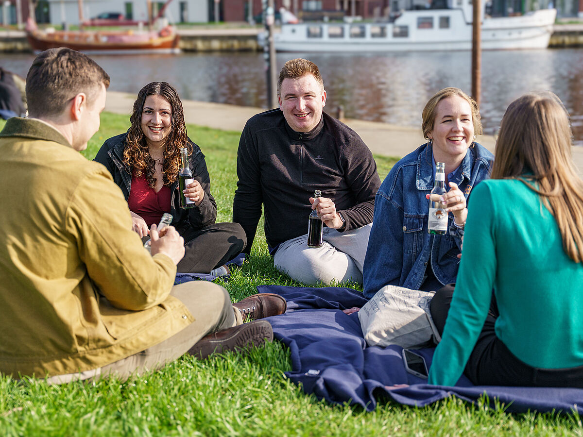 Fünf Studierende sitzen auf dem Rasen am Ryck, unterhalten sich und halten Erfrischungsgetränke in der Hand.