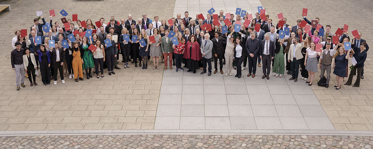 Viele Menschen stehen für ein Gruppenbild im Innenhof einer alten Universität und halten Abschlussurkunden in die Luft.