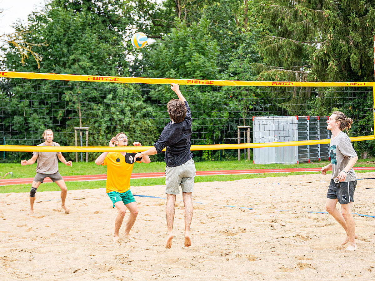 Eine Gruppe von 4 jüngen Männern sind sommerlich gekleidet und spielen auf einem sandigen Platz Beachvolleyball. Im Hintergrund stehen grüne Bäume.
