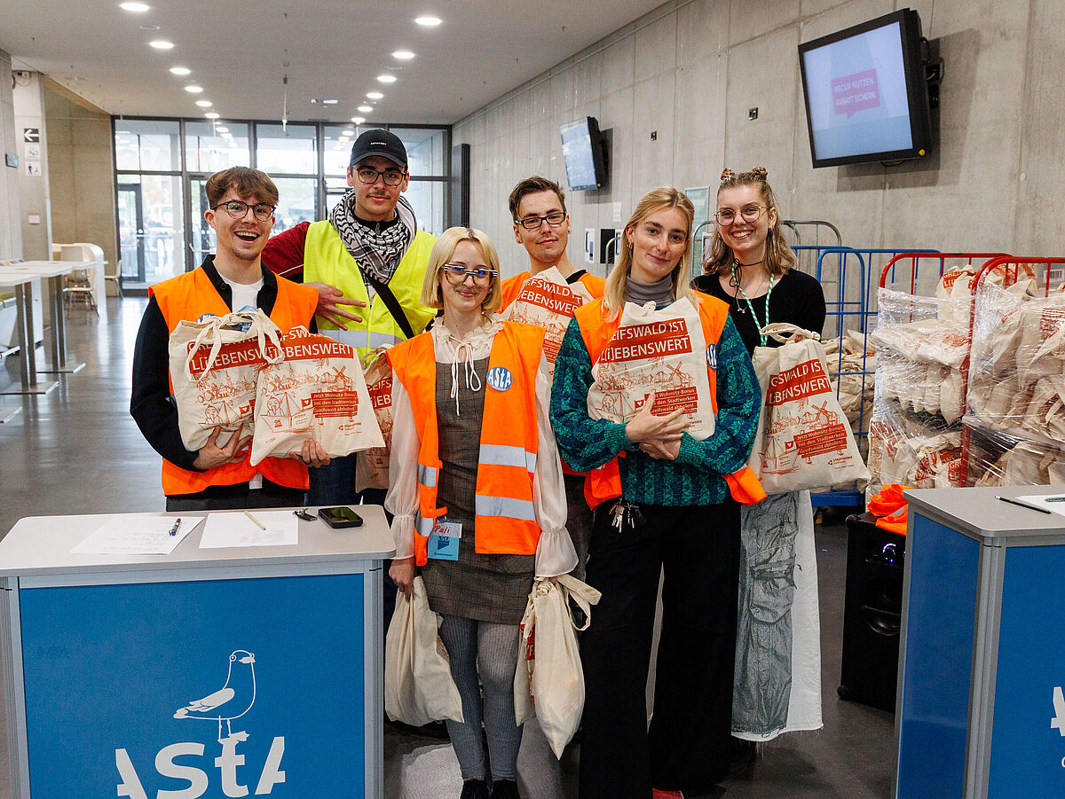 Eine lächelnde Gruppe freundlicher Studierender trägt neonorangefarbene Westen und steht hinter einem Aufsteller des AStA.